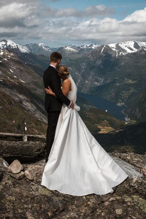 A couple in wedding attire embrace while standing on a mountain overlook with snow-capped peaks and valleys in distance.