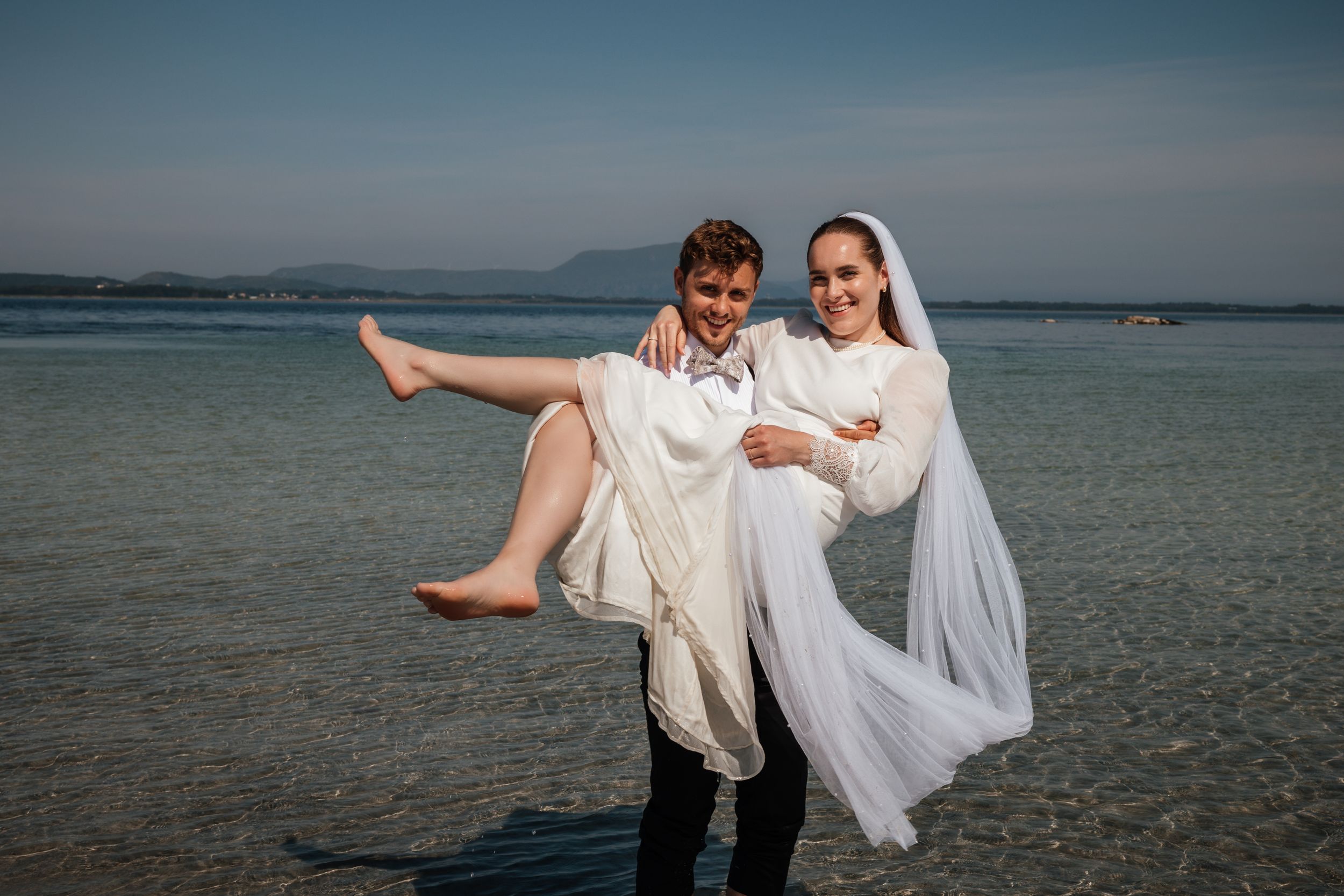 A couple in wedding attire stands at the beach with calm blue water and mountains in the distance.