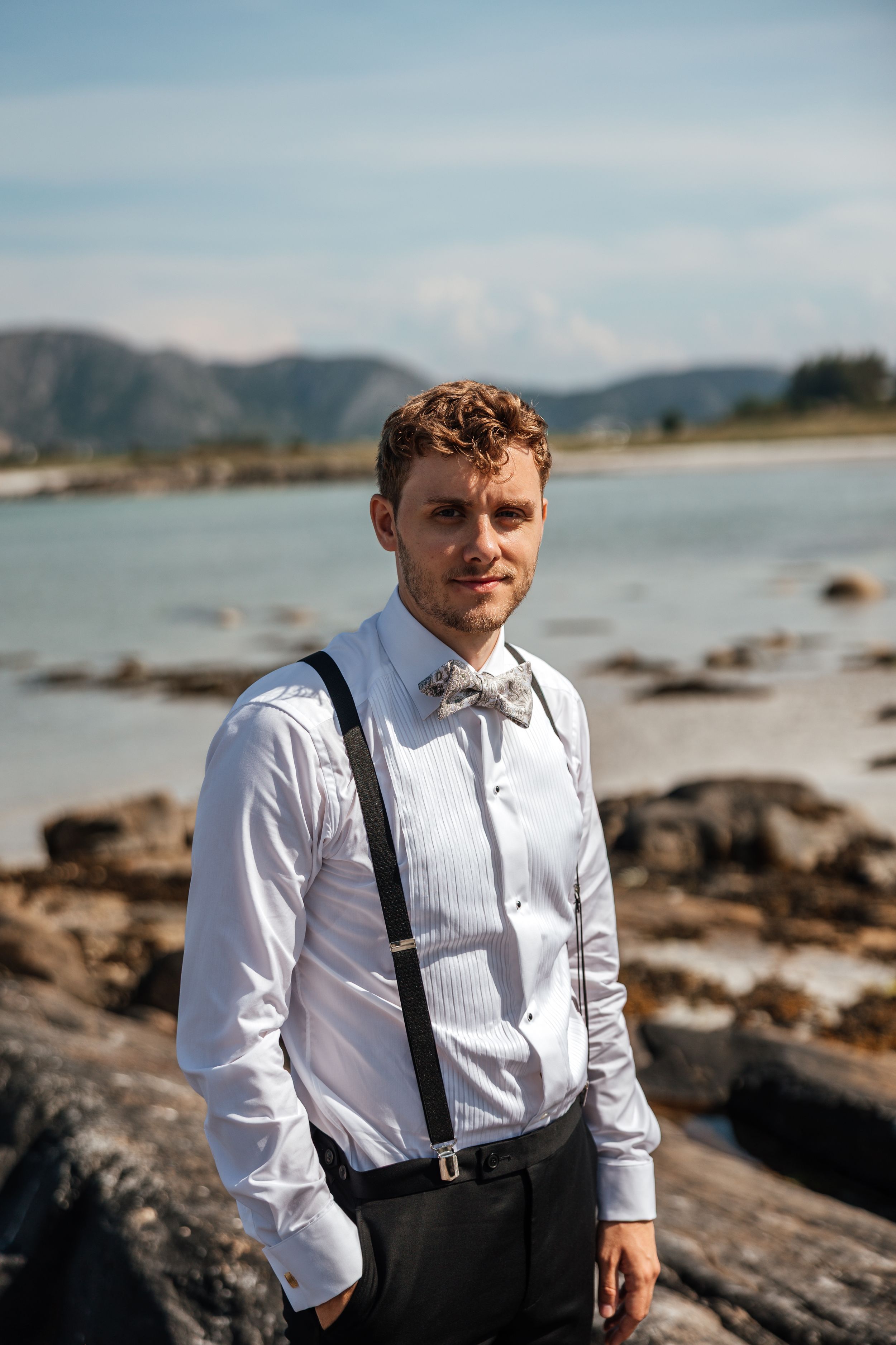Someone in formal attire with suspenders and bow tie poses along a rocky coastline with mountains in the background.