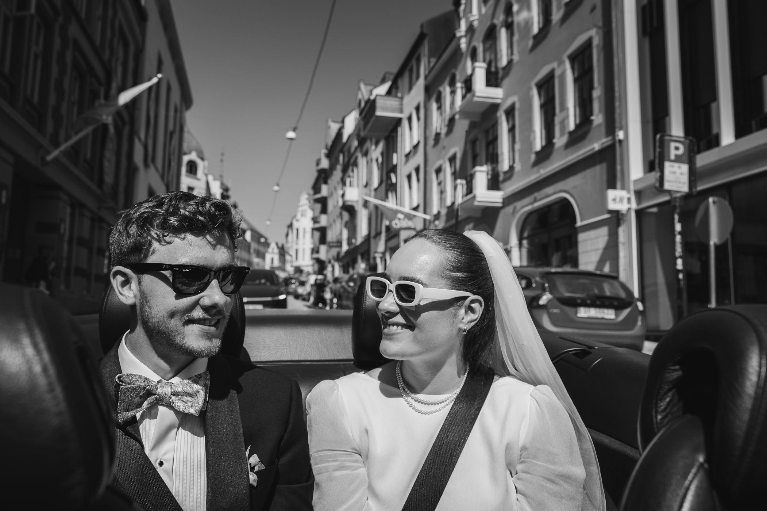 A couple wearing sunglasses and formal attire walk together on a sunny European street lined with historic buildings.