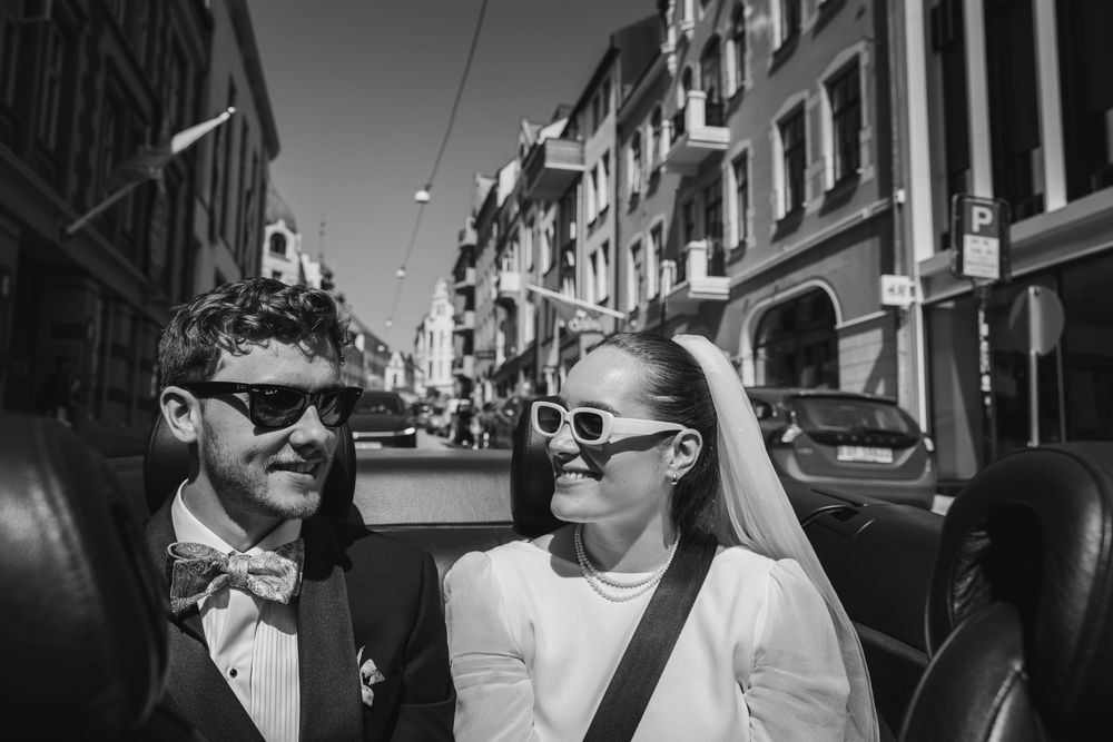 A couple wearing sunglasses and formal attire walk together on a sunny European street lined with historic buildings.