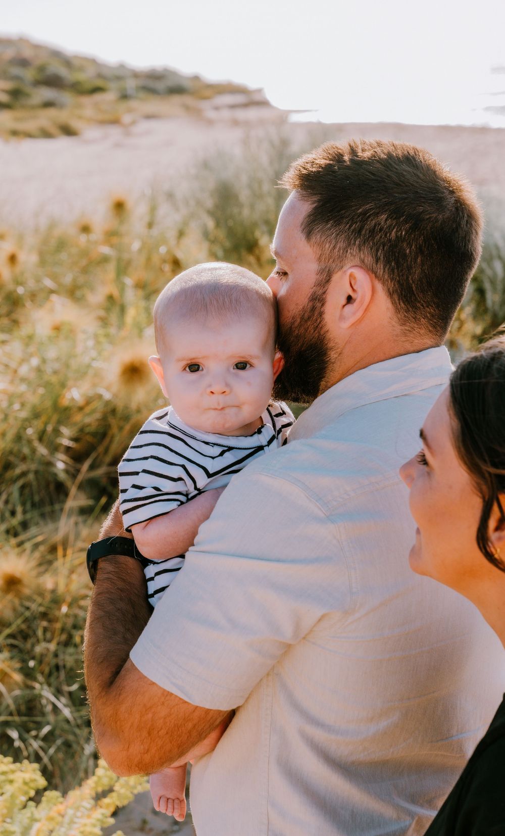 Tender moments between parent and baby in striped outfit against coastal backdrop.