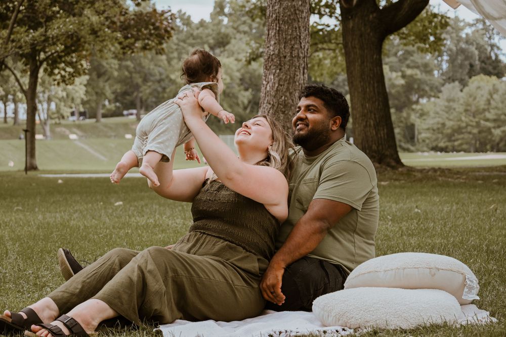 Happy interracial family sitting on grass in park with parents lifting smiling baby in the air.