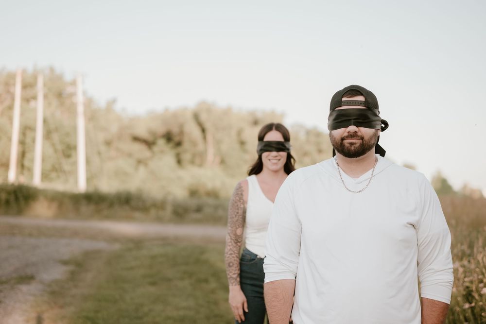 A couple stands on a rural path, man in white shirt and black cap in foreground, woman behind.