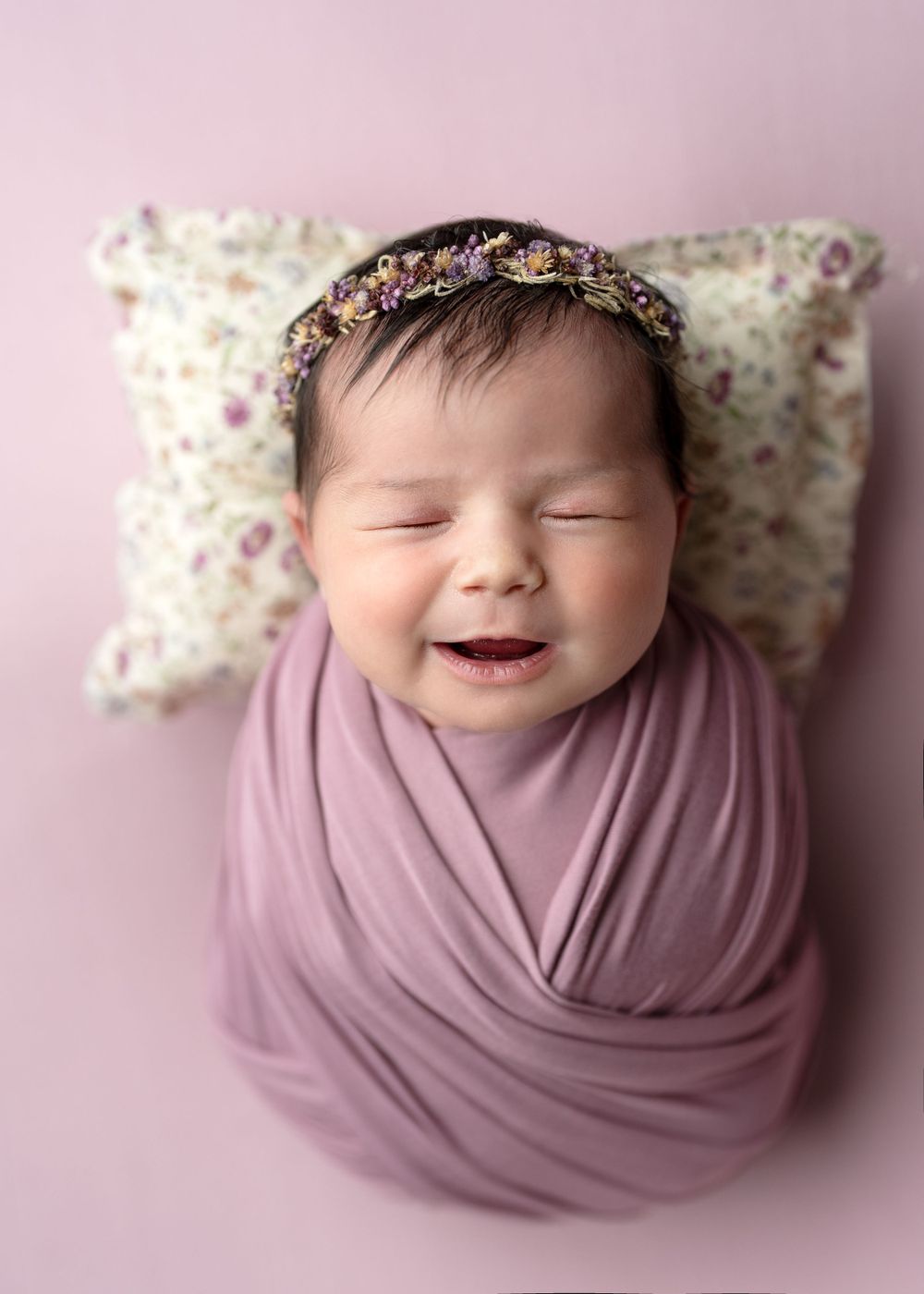 Newborn wrapped in mauve fabric smiling with floral headband against pink background.