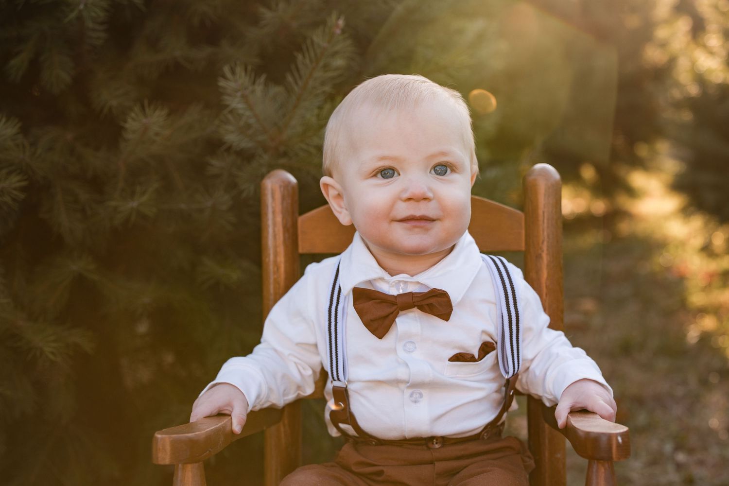 Small baby wearing bow tie and suspenders sits in wooden chair during golden hour outdoor photo session.