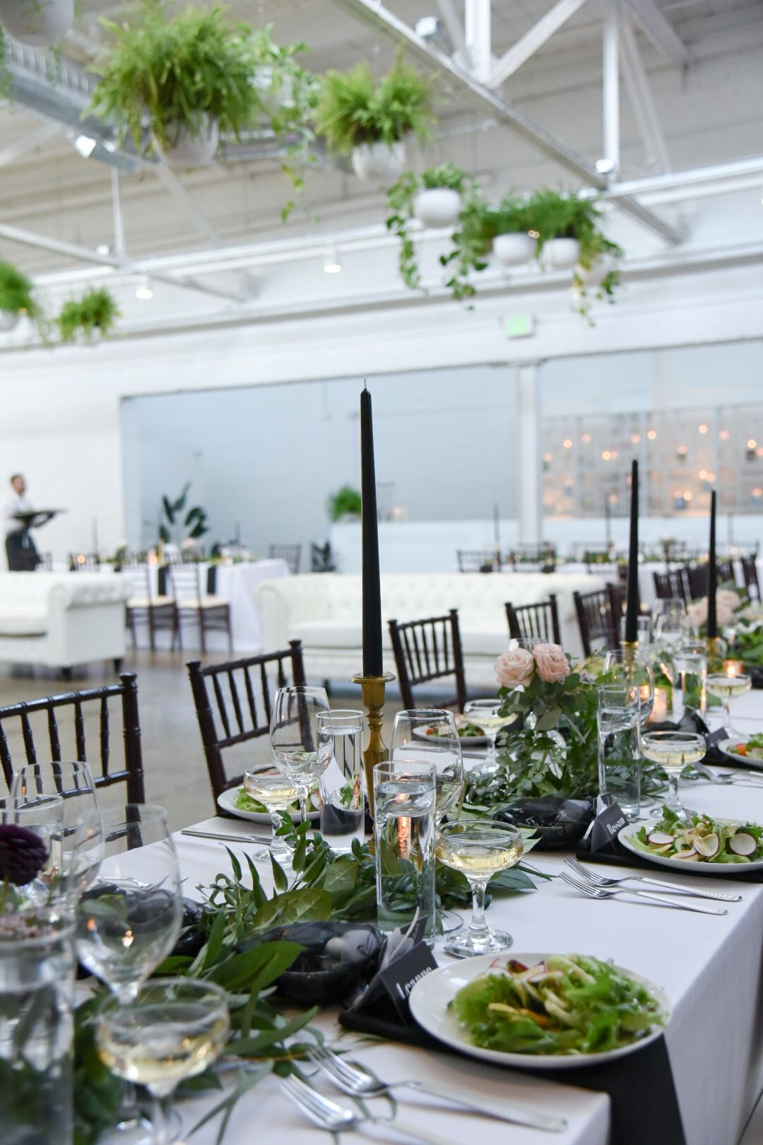 Elegant wedding reception tables set with white linens, black chairs, and hanging greenery in an industrial-style venue.