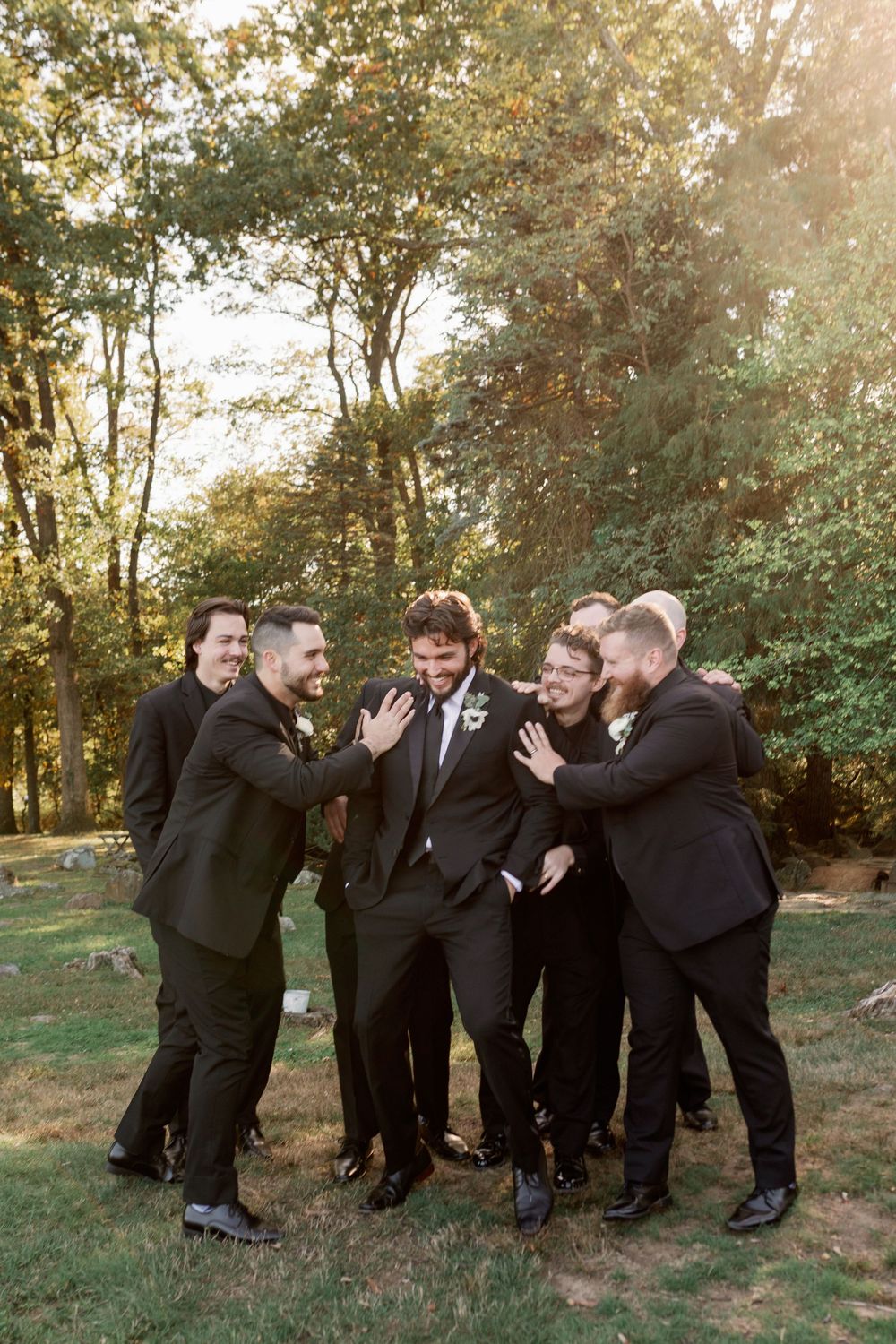 Groomsmen in black suits laugh and gather around in a playful moment outdoors.