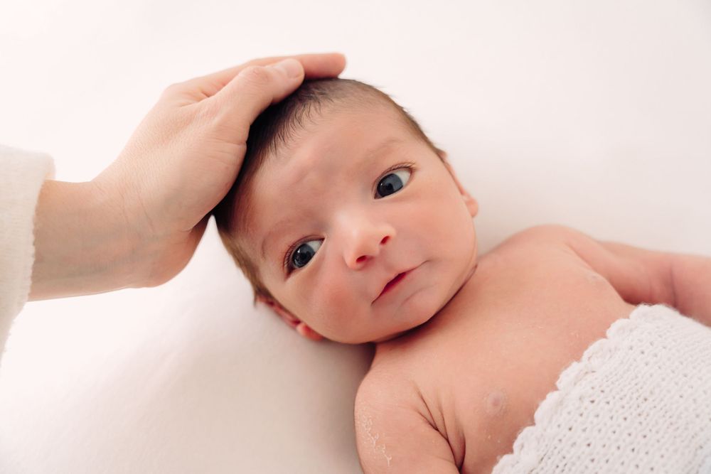 Gentle hand resting on newborn baby's head wrapped in white blanket during portrait session.