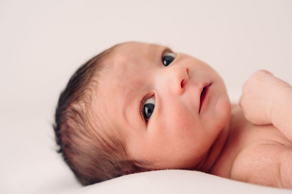 Close-up portrait of an alert newborn baby with dark hair against white background.