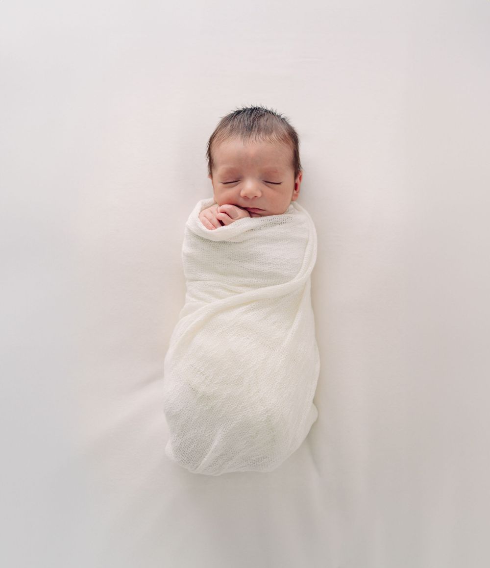 Peaceful newborn baby swaddled in white fabric against light background.