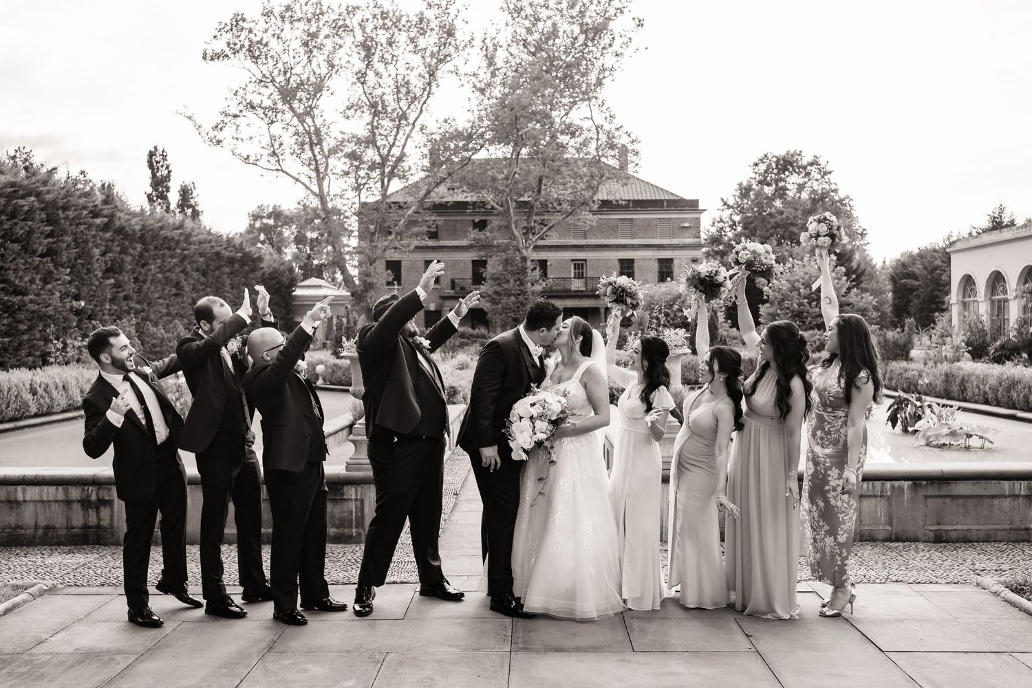 A wedding party poses outdoors on a stone terrace with trees and ornate architecture in the background, black and white photo.