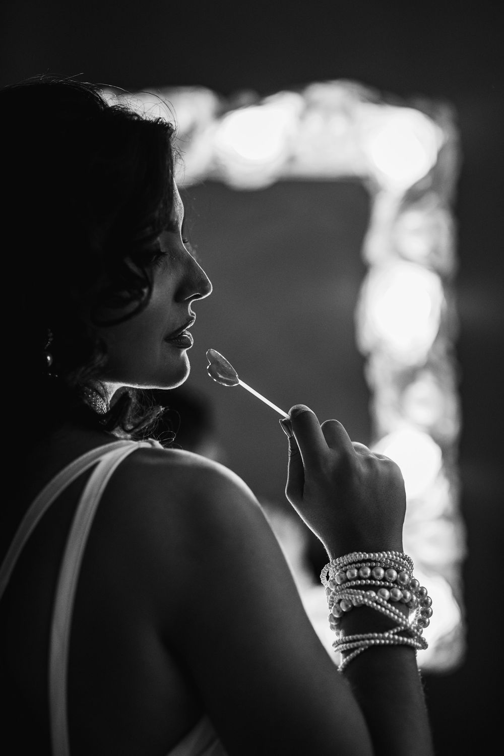 Black and white photo of a woman applying lipstick, wearing pearl bracelets, with a mirror in the background.