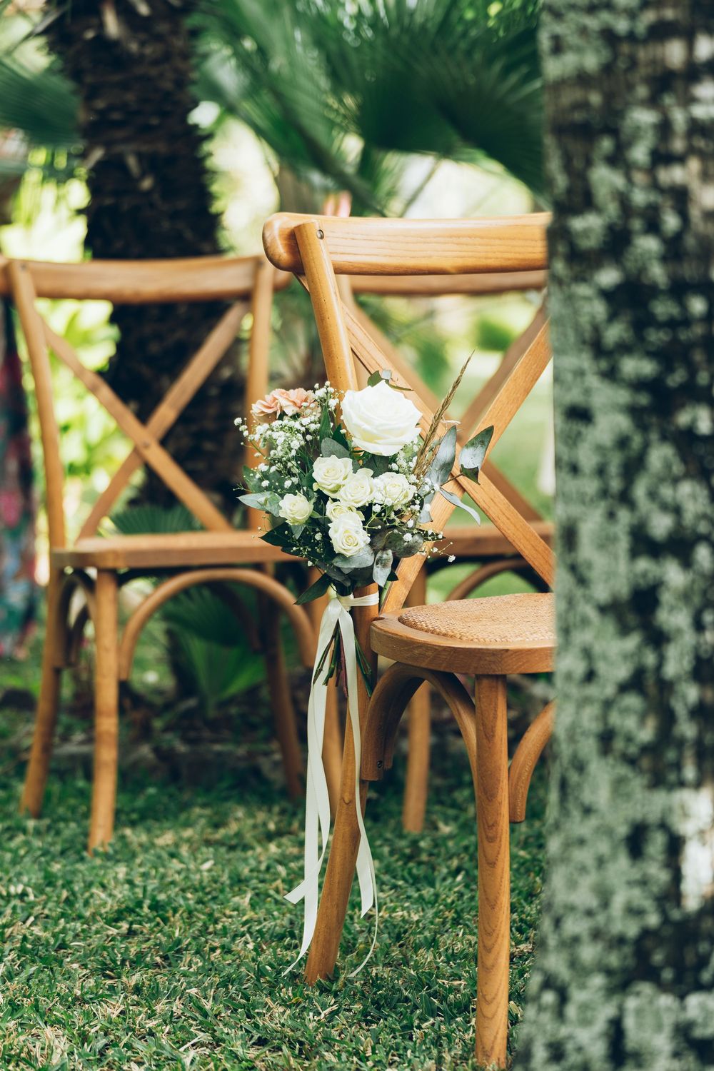 Wooden cross-back chairs decorated with white roses and ribbon bouquets on green grass at an outdoor wedding ceremony.