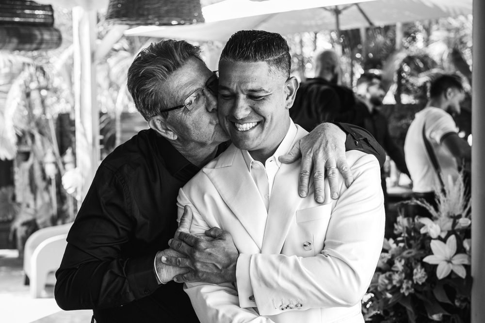 Two men embracing and smiling in a black and white photo at an outdoor event, one kissing the other on the cheek.