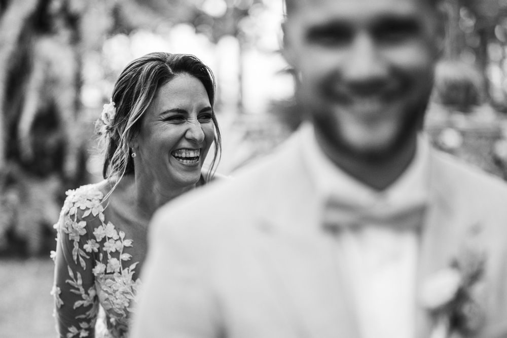 Black and white photo of a laughing bride in floral dress with groom in bow tie at an outdoor wedding.