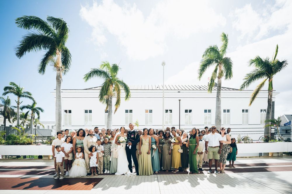 A large wedding party poses together outdoors under towering palm trees in front of a white building on a sunny day.