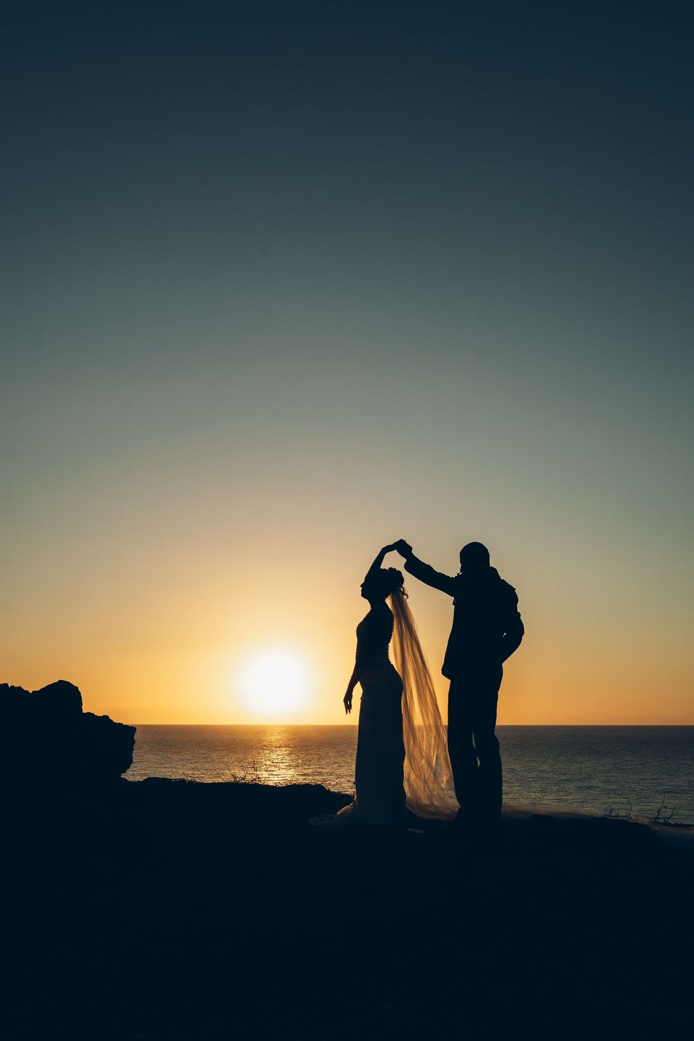 Silhouetted couple dancing at sunset on a beach with golden sun reflecting off the ocean water.