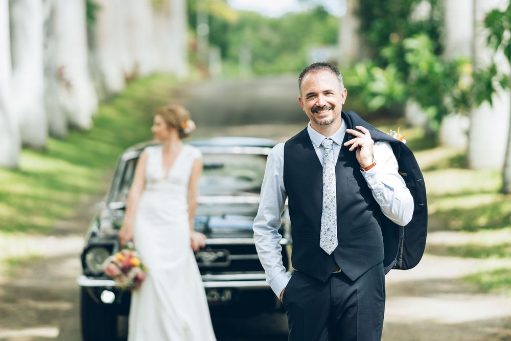 Wedding couple poses on a tree-lined street with a classic black car in the background.