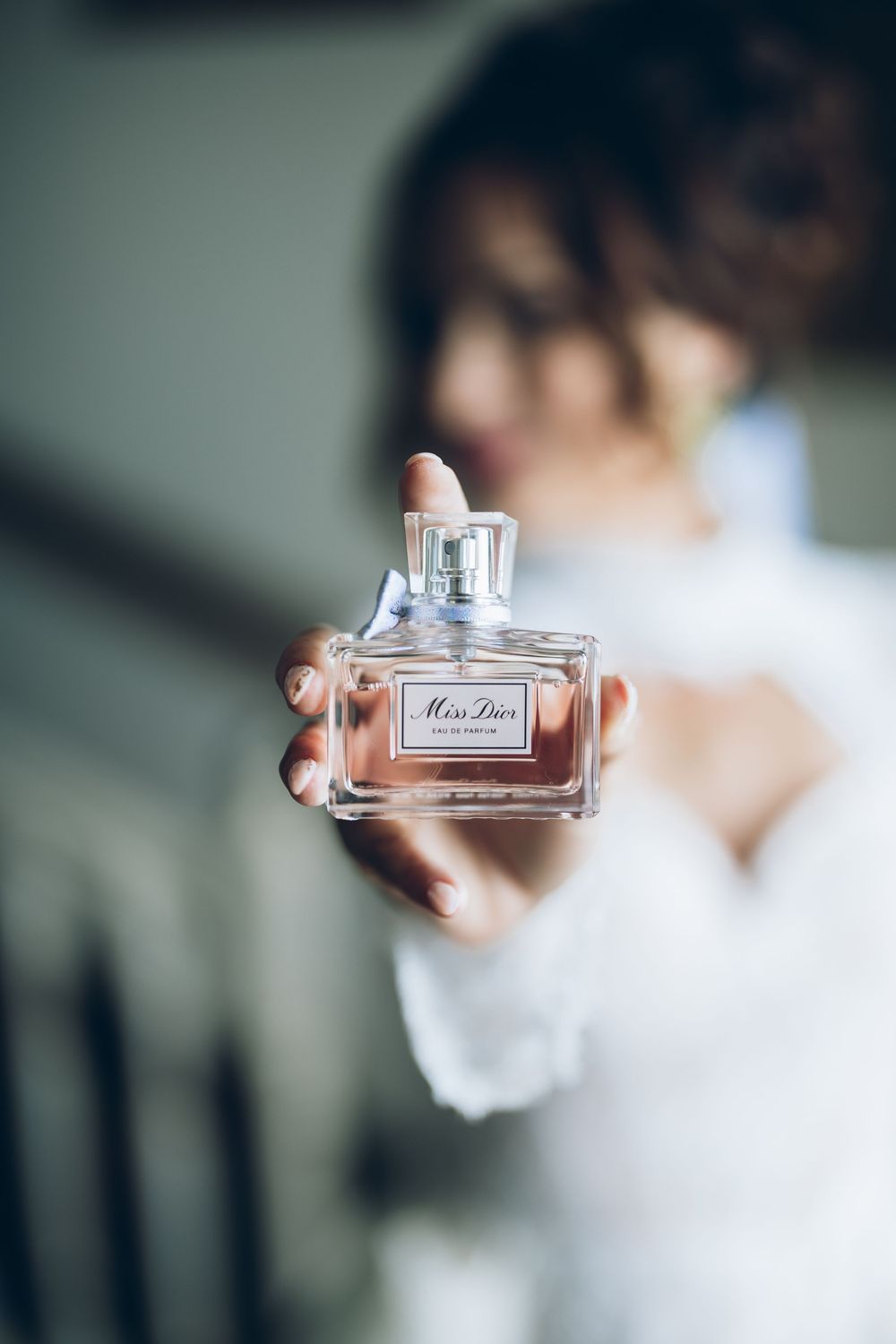 A bottle of Miss Dior perfume being held up against a soft, blurred background in natural light.