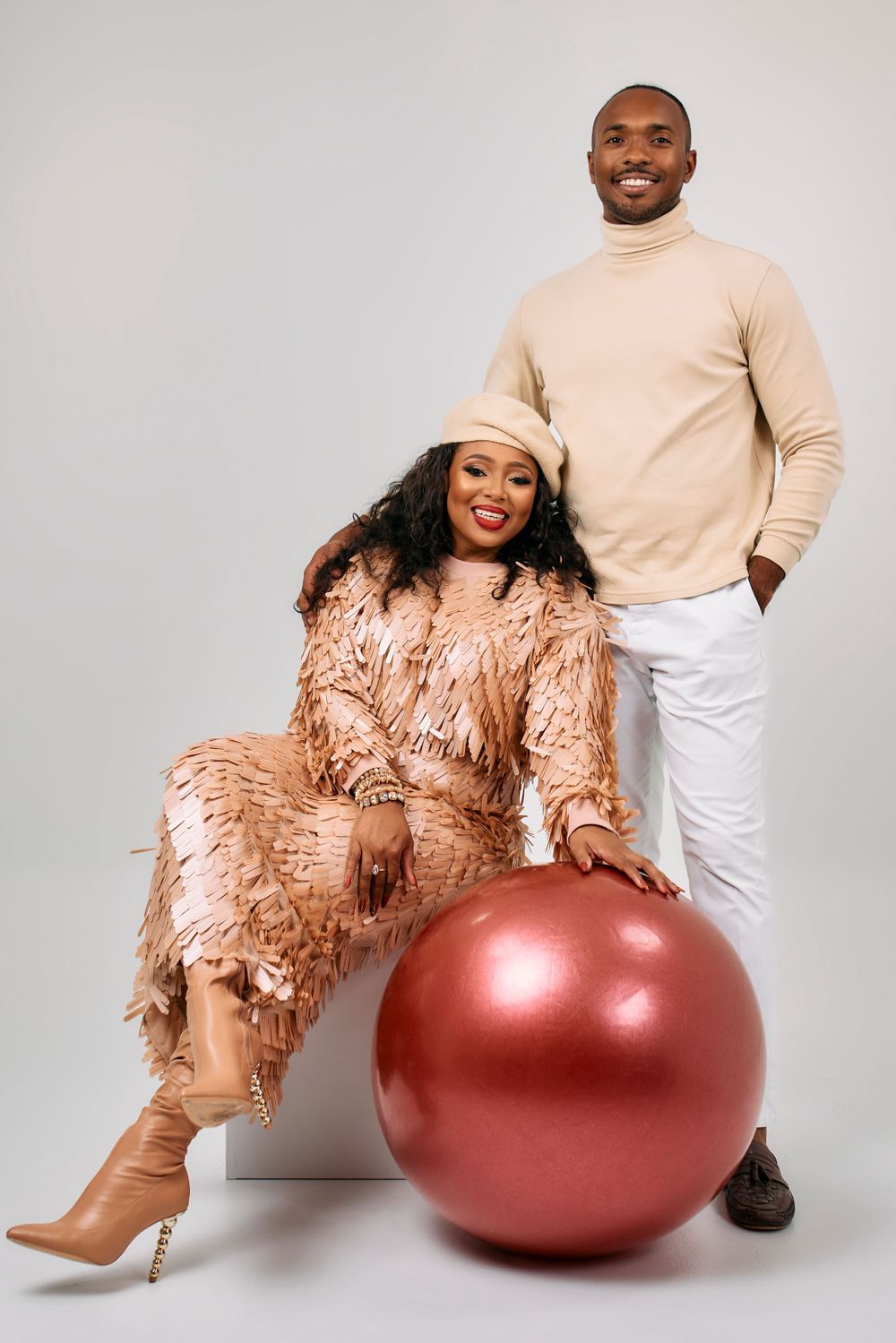 Studio portrait with person in beige turtleneck standing while other sits on large red exercise ball.
