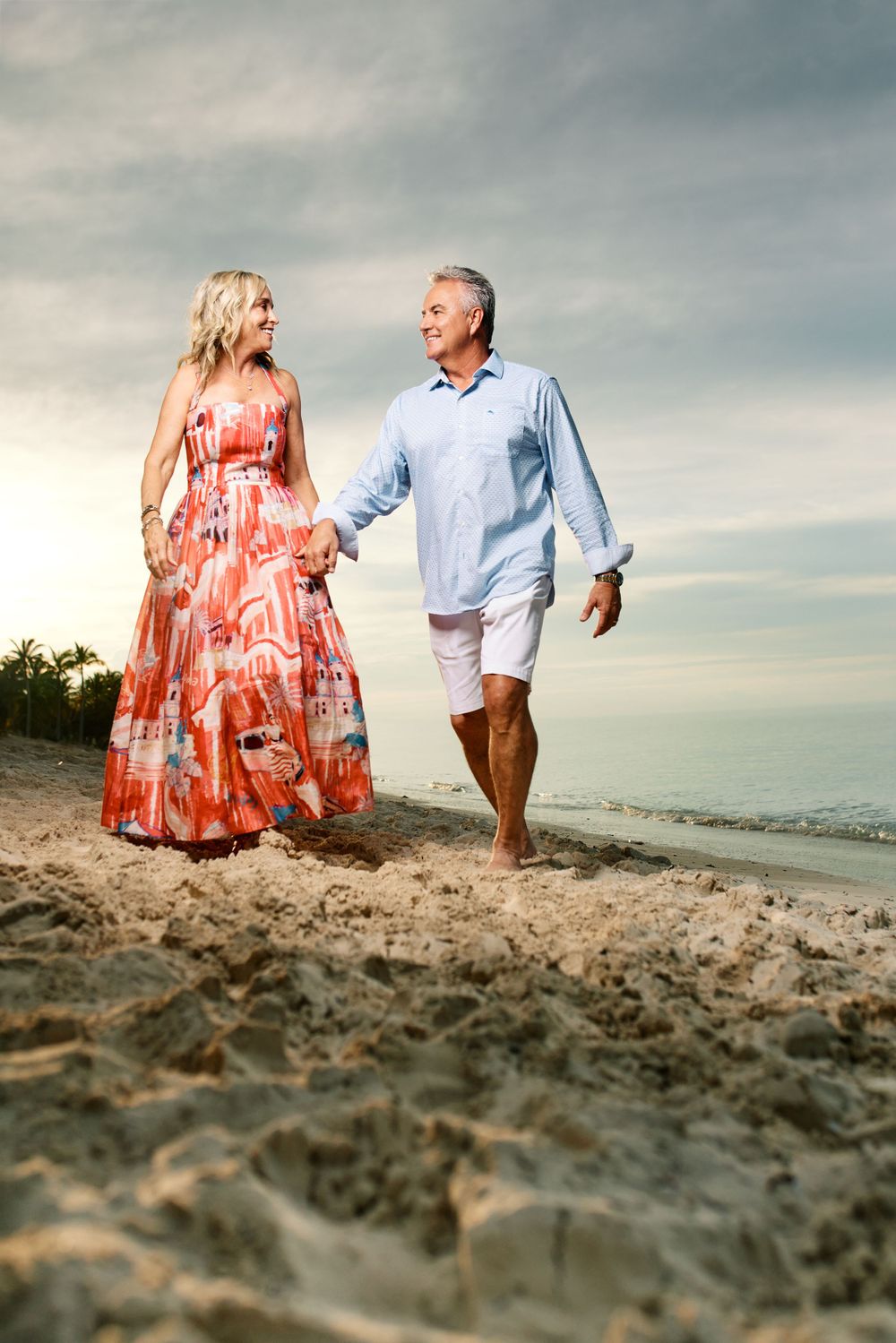 Couple walking on beach at sunset with woman in coral maxi dress and man in light blue shirt.
