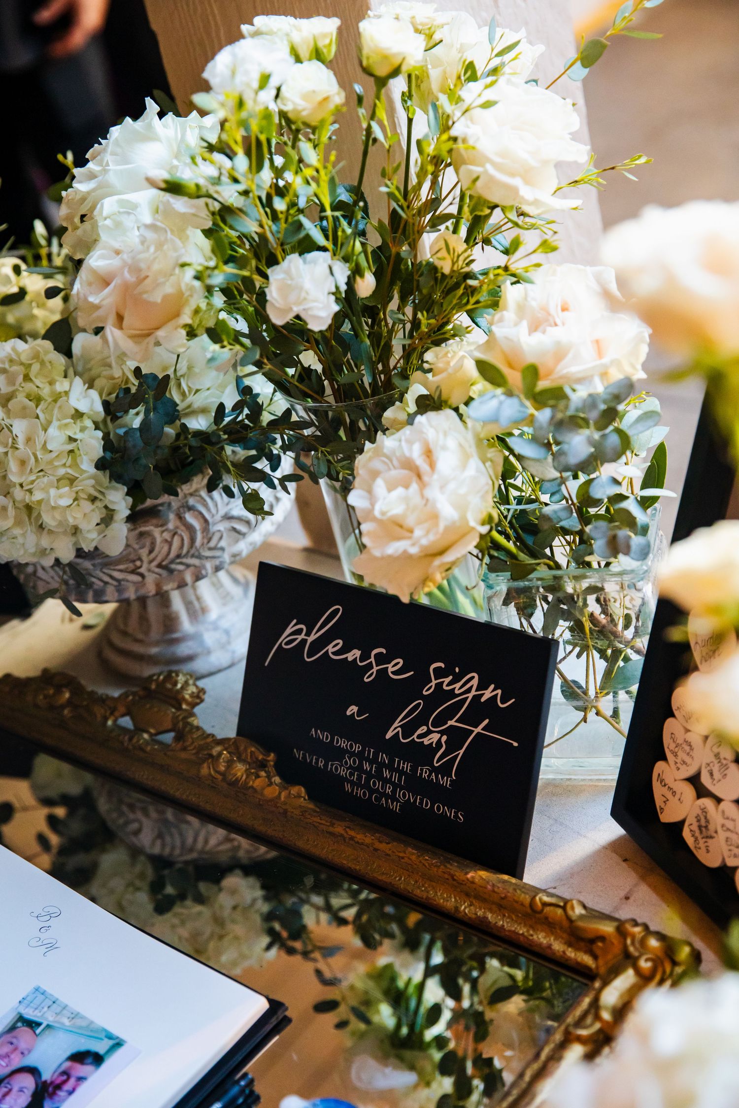 White roses and floral arrangements adorn a sign-in area with a black sign reading 'Please sign our guest' at an event.