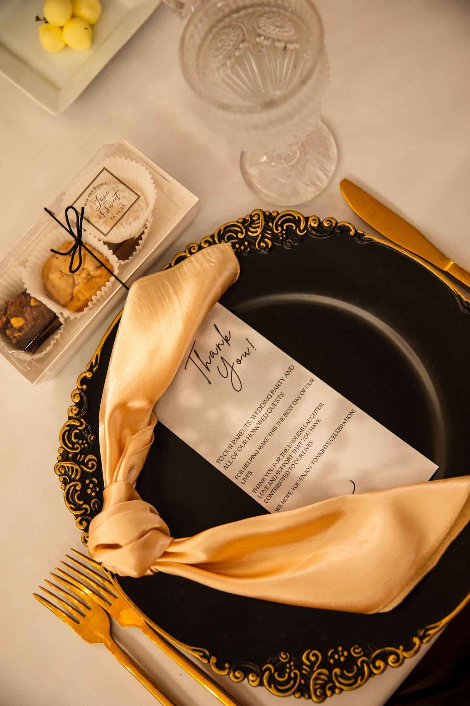 Elegant place setting with black and gold plate, champagne napkin, gold cutlery and menu card on cream tablecloth.