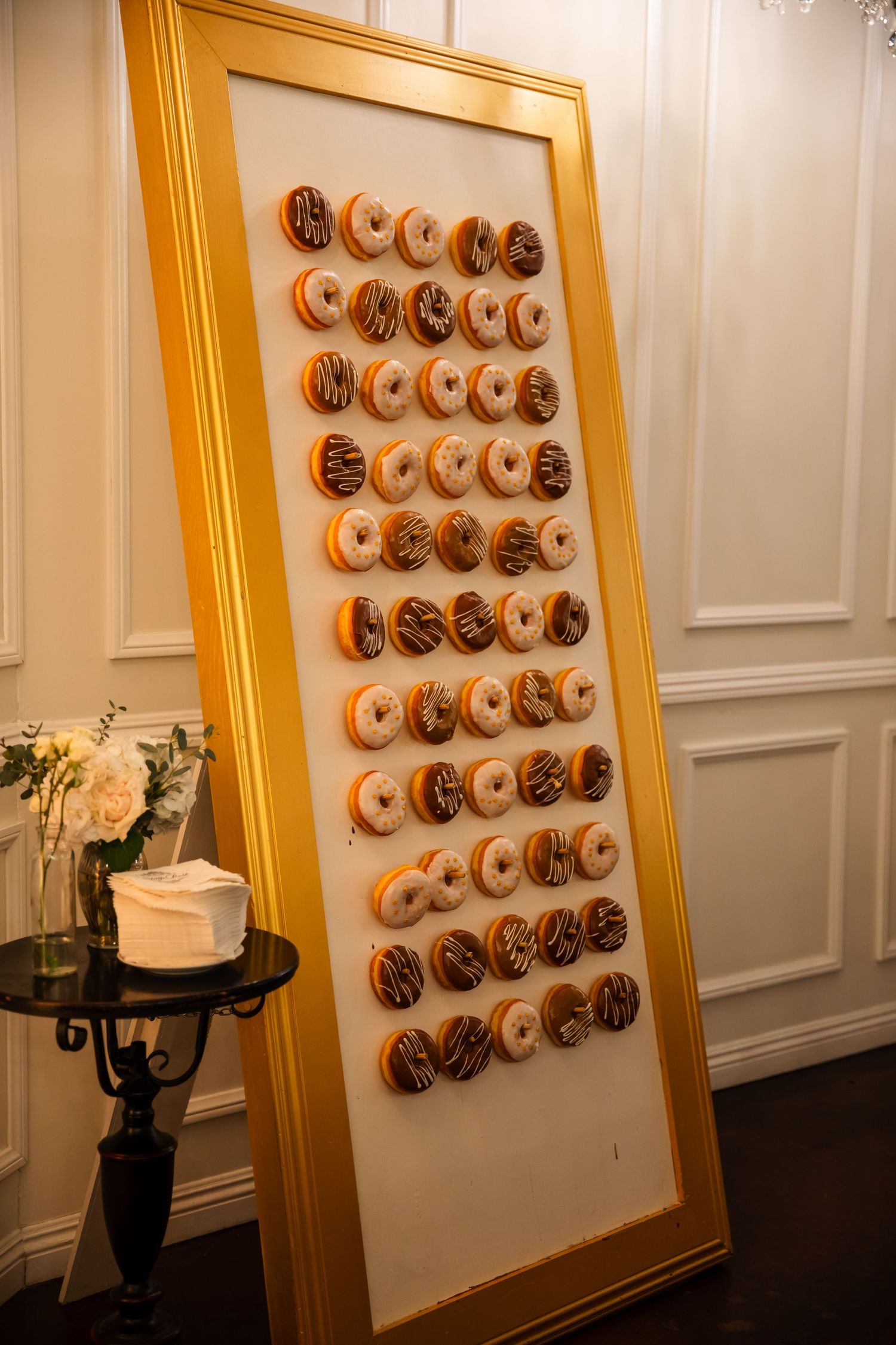 A gold-framed wall display featuring rows of decorated donuts in chocolate and vanilla frosting against a white backdrop.