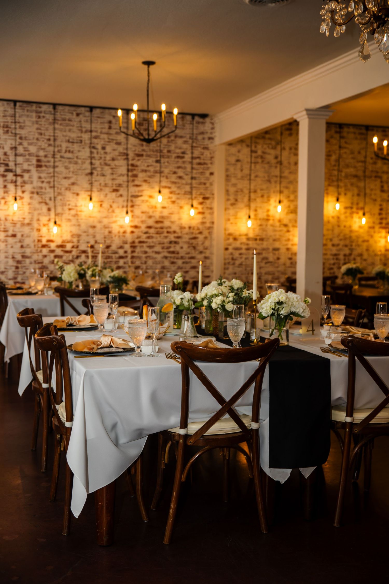 Elegant wedding reception table setup with white linens and floral centerpieces against exposed brick wall with warm lighting.
