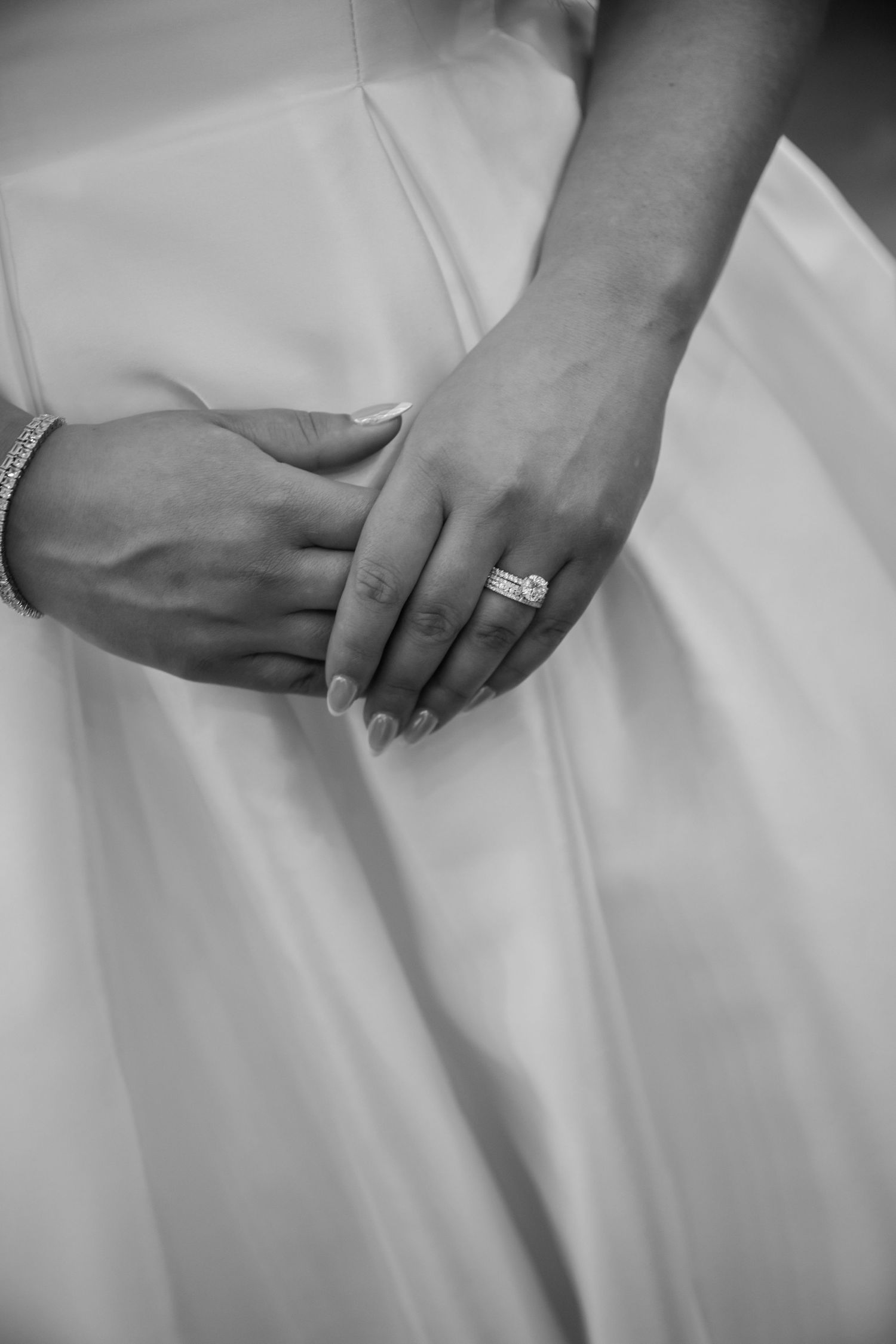 Black and white detail shot of hands holding white wedding dress fabric, featuring an engagement ring and bracelet.