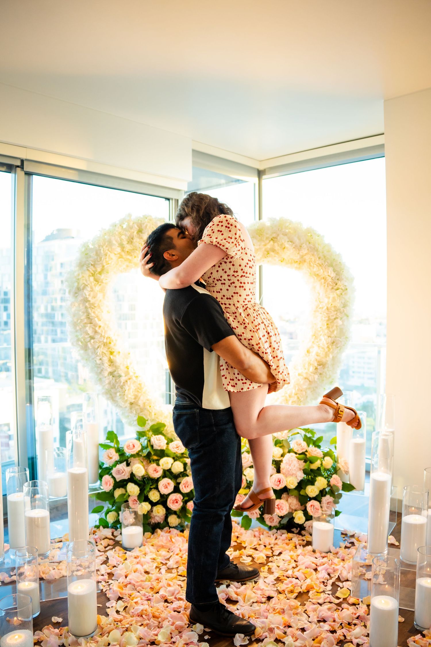 A romantic proposal sequence in a room decorated with pink rose petals and a heart-shaped floral backdrop.