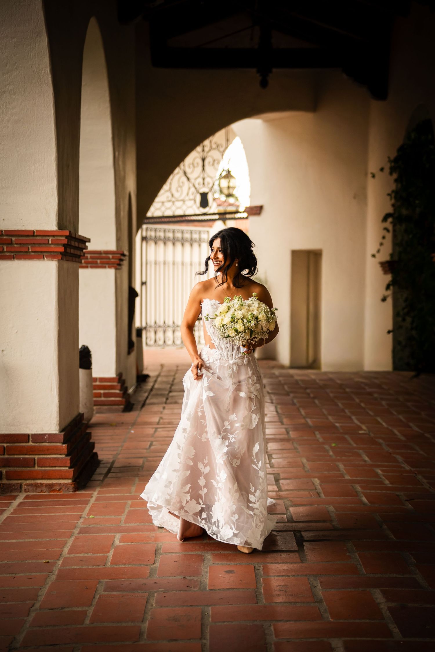 A bride in a flowing white lace wedding dress walks along a Spanish-style corridor with terracotta tile floors.