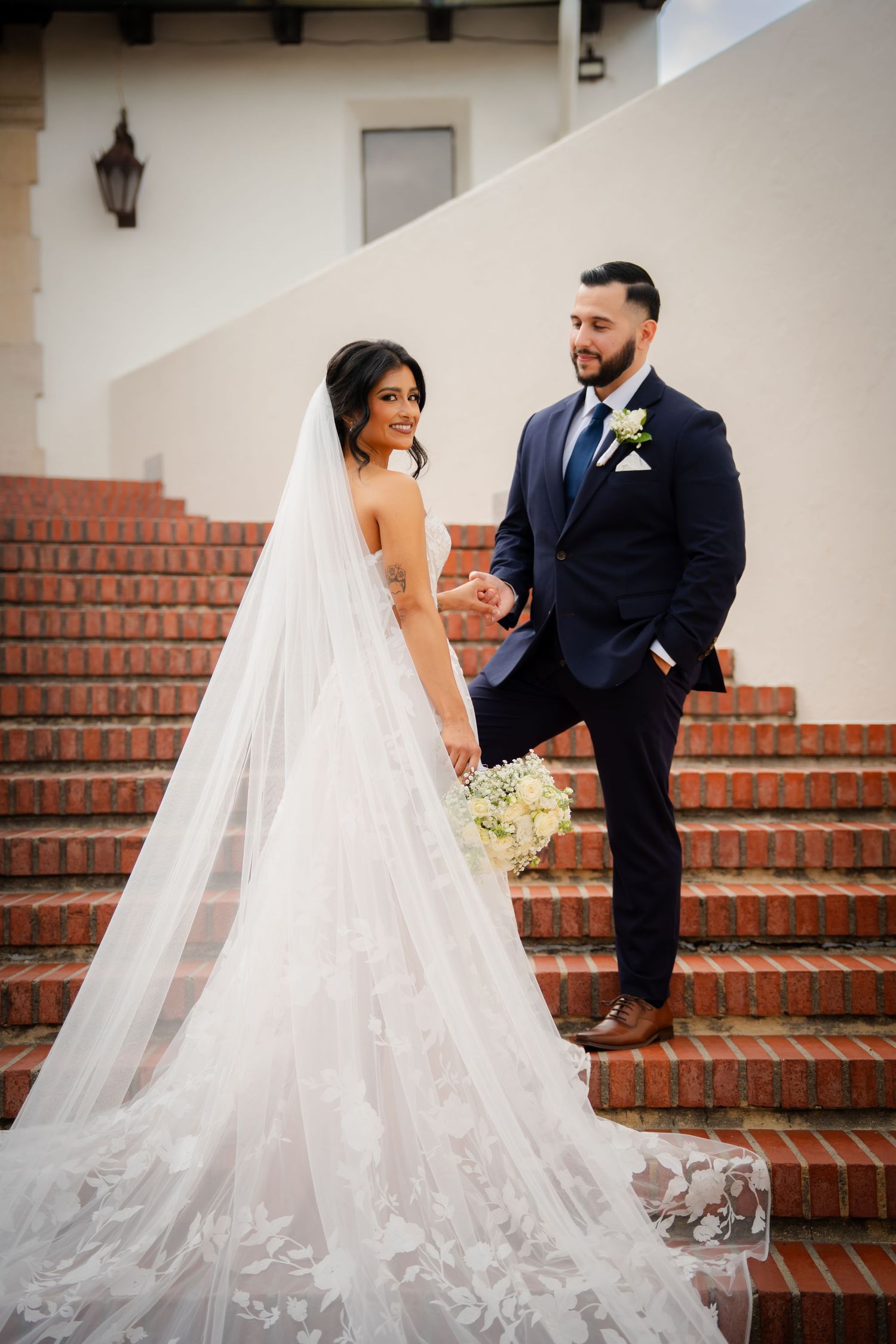 A romantic wedding scene on brick steps with a flowing white veil and navy suit.