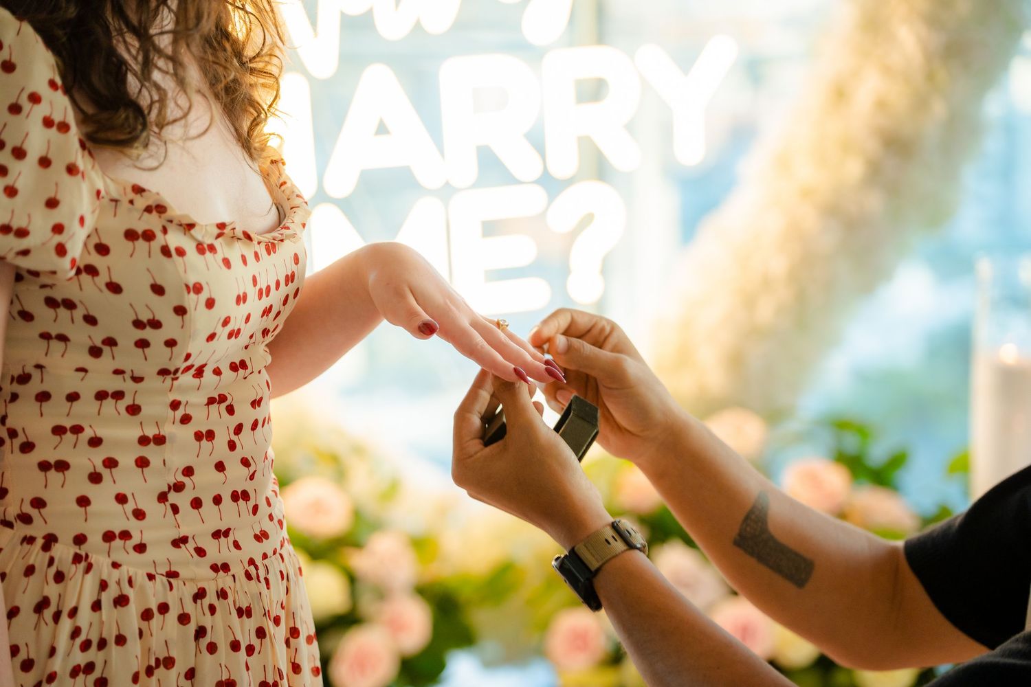 A person in a cherry-print dress receives a ring during an outdoor proposal with sunlight streaming through the trees.