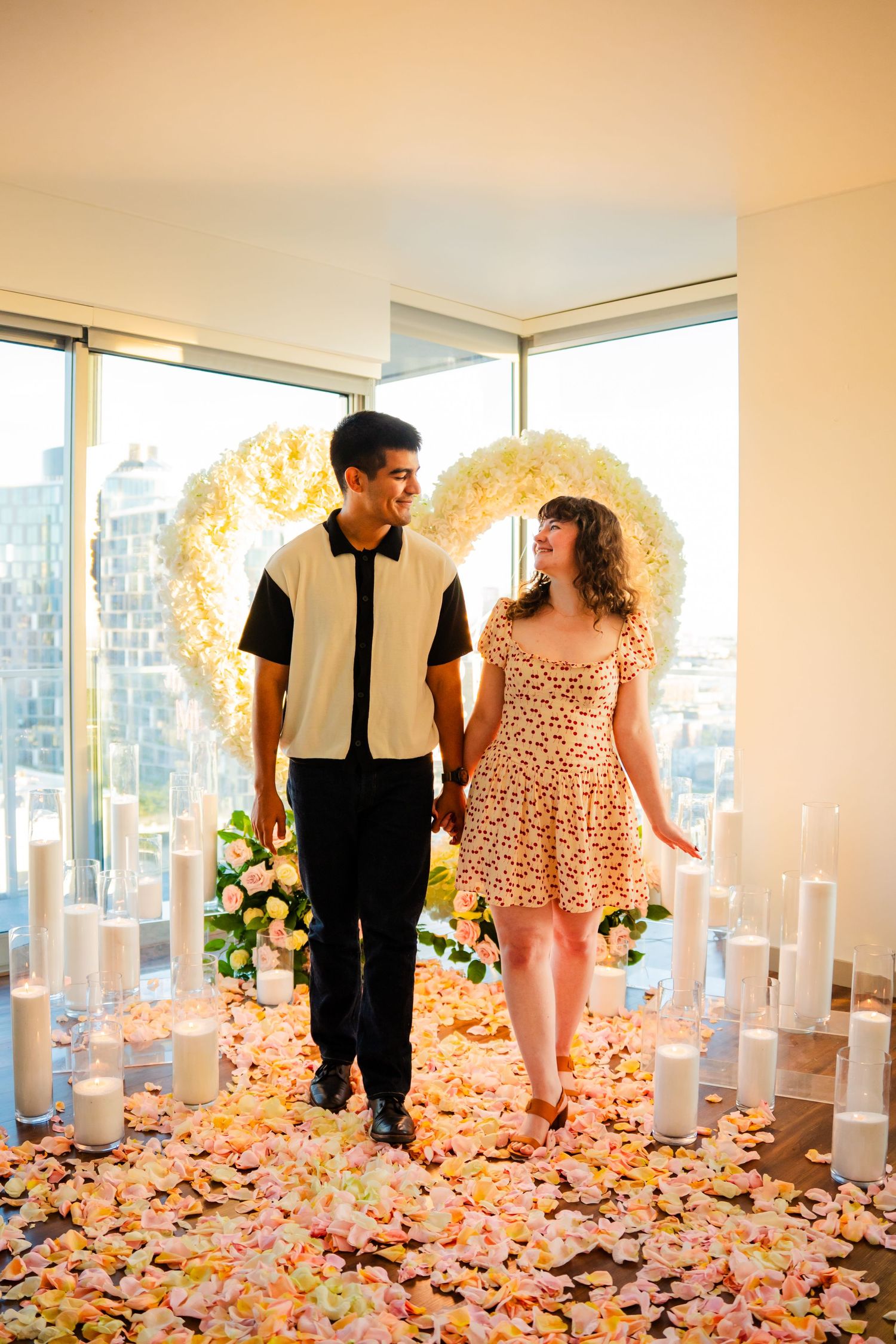A couple stands in a romantic setting with flower petals on the floor, candles and a floral arch in a high-rise apartment.