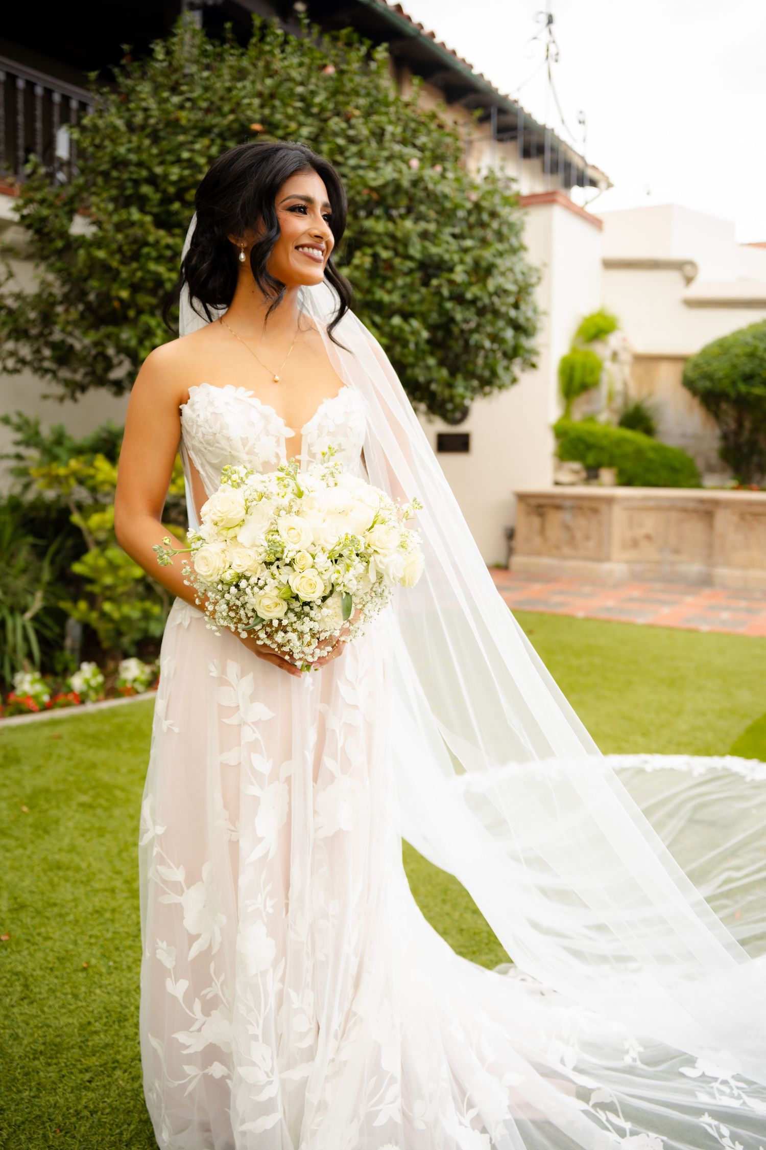 A bride in a delicate lace wedding gown poses in a garden setting with a flowing veil.
