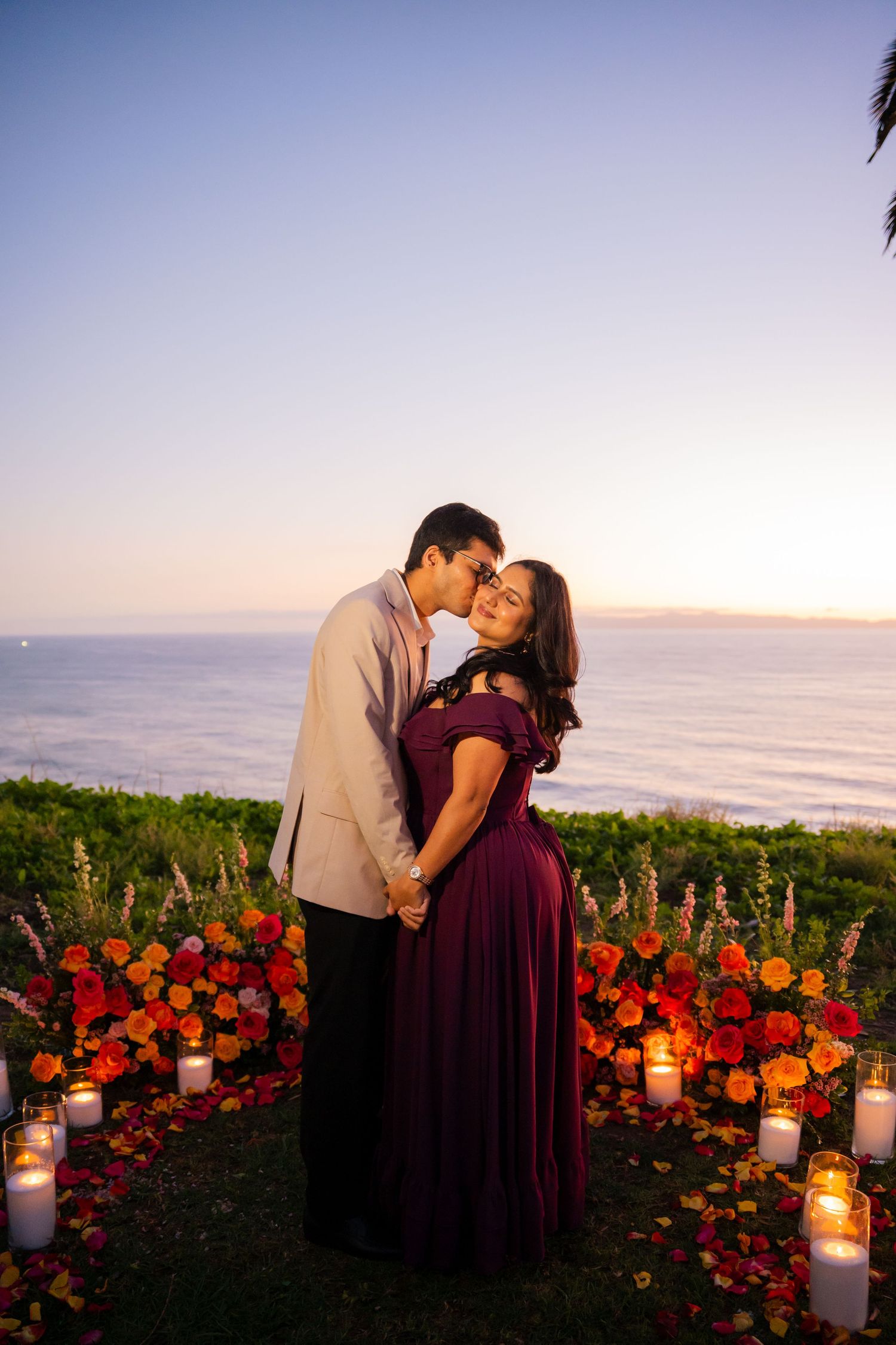 A romantic couple embraces at sunset by the ocean with glowing candles and vibrant red flowers in the foreground.