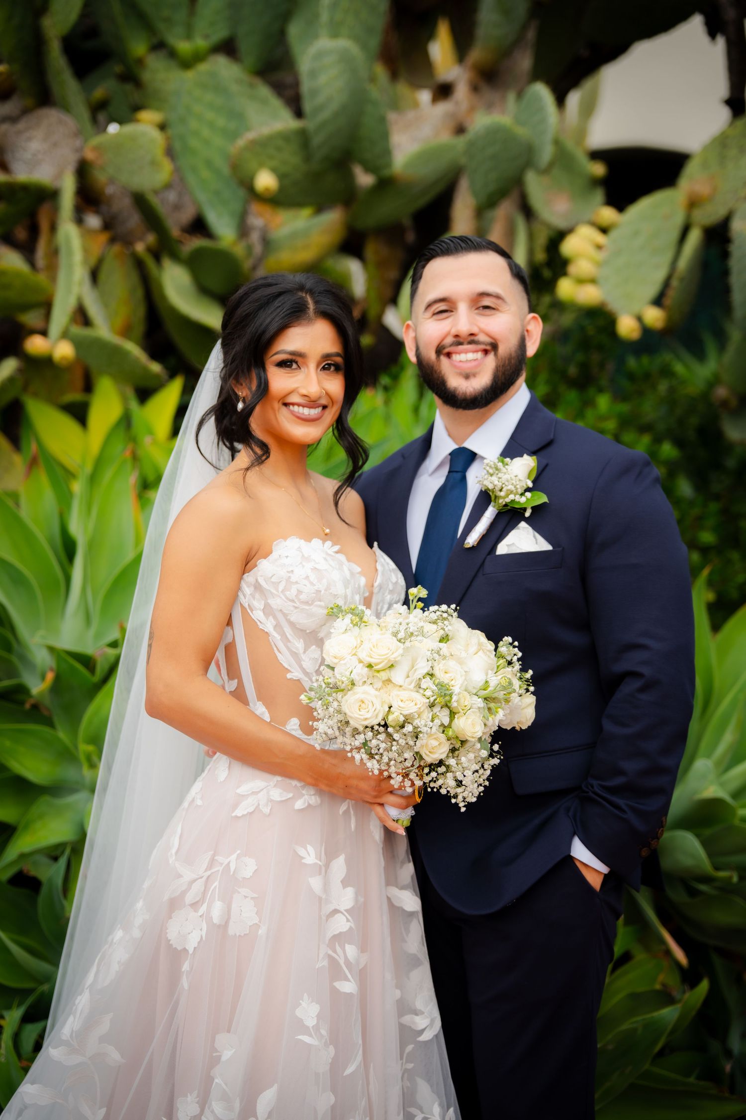 Happy newlyweds pose together in a lush garden setting with white floral bouquet.