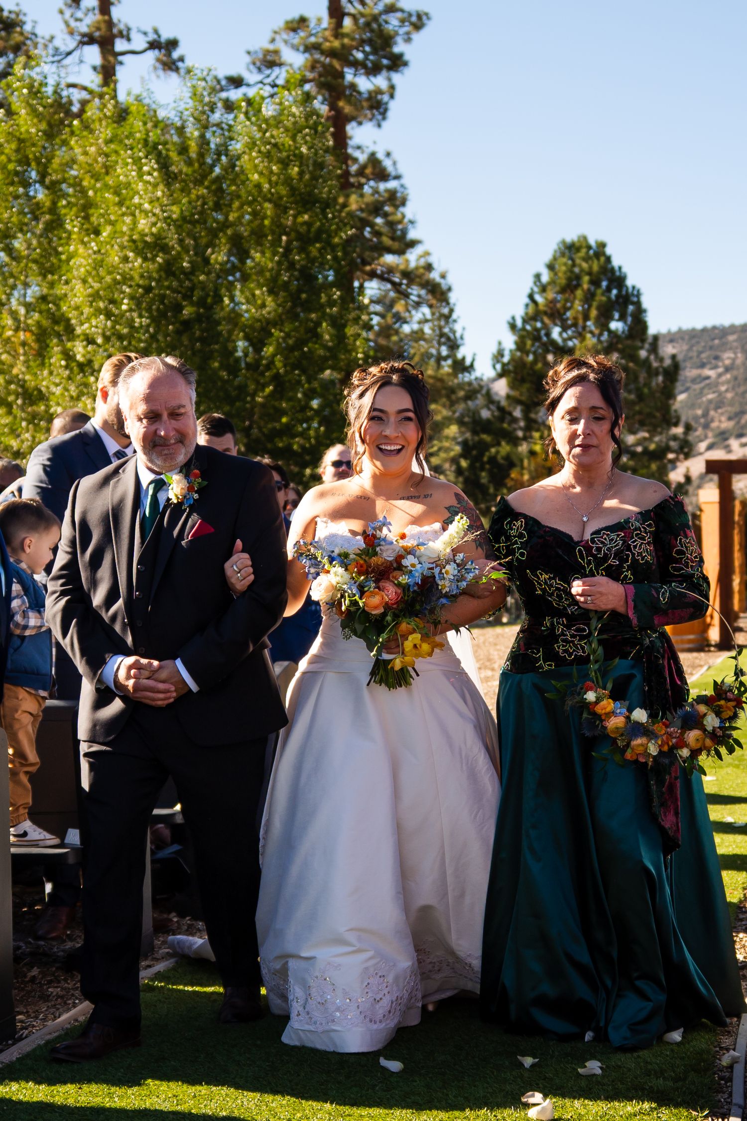 Wedding party members laugh together outdoors on a sunny day while standing on green grass with trees in background.