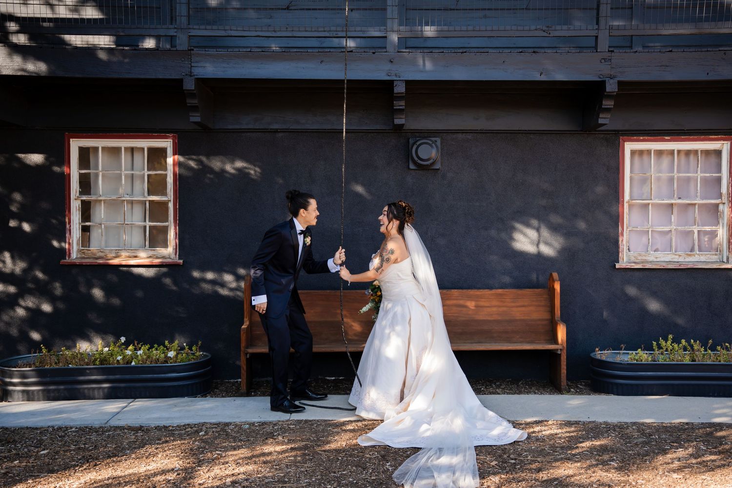 A couple in wedding attire sits together on a wooden bench outside a dark building with white windows.