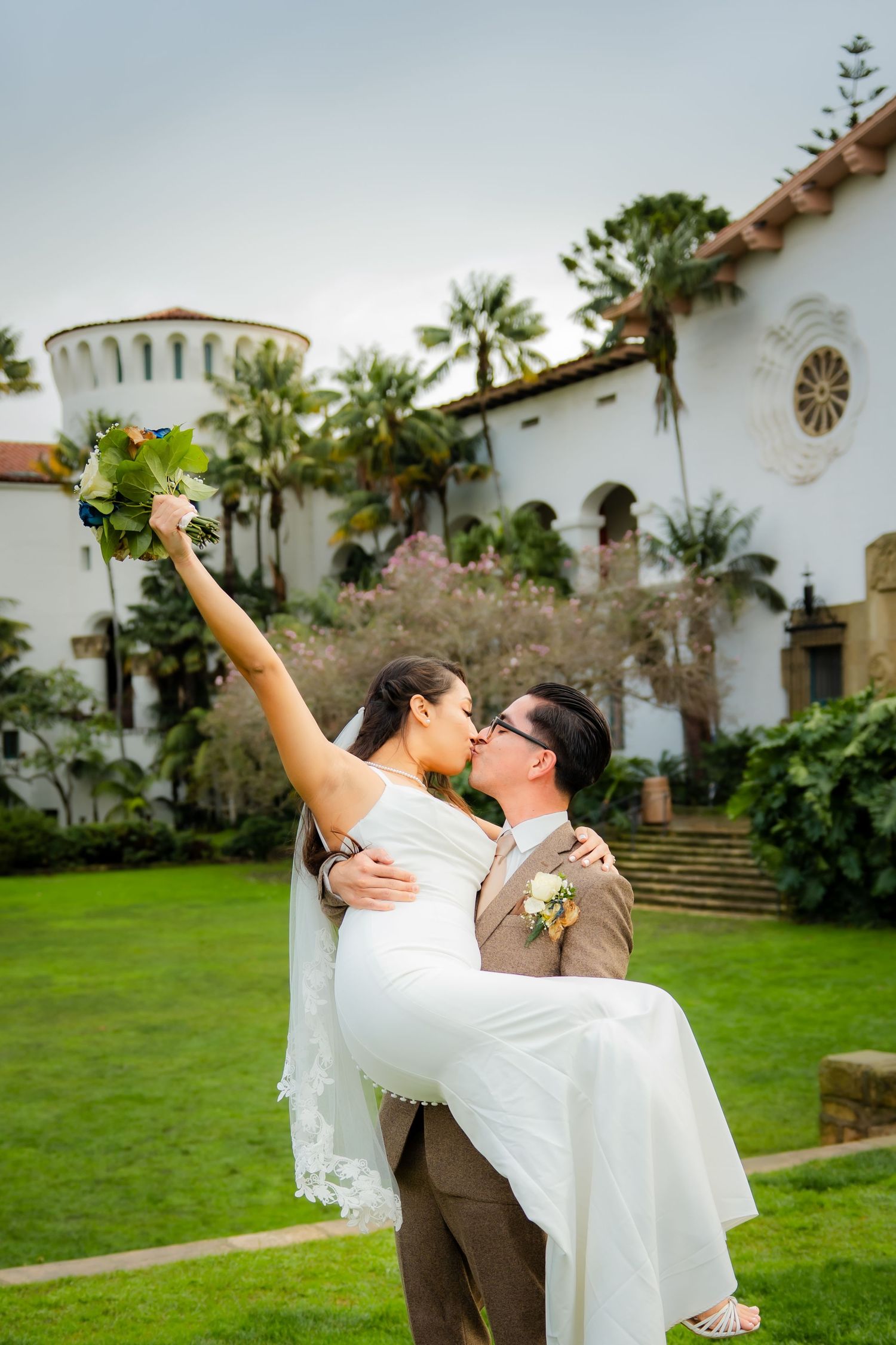 A couple in wedding attire dances joyfully on a green lawn in front of a Spanish-style building with white walls.