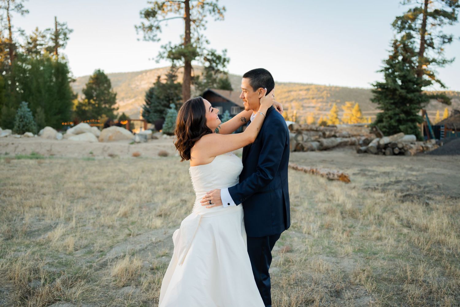 A couple shares an intimate dance moment on a grassy hillside during golden hour with tall pine trees in the background.