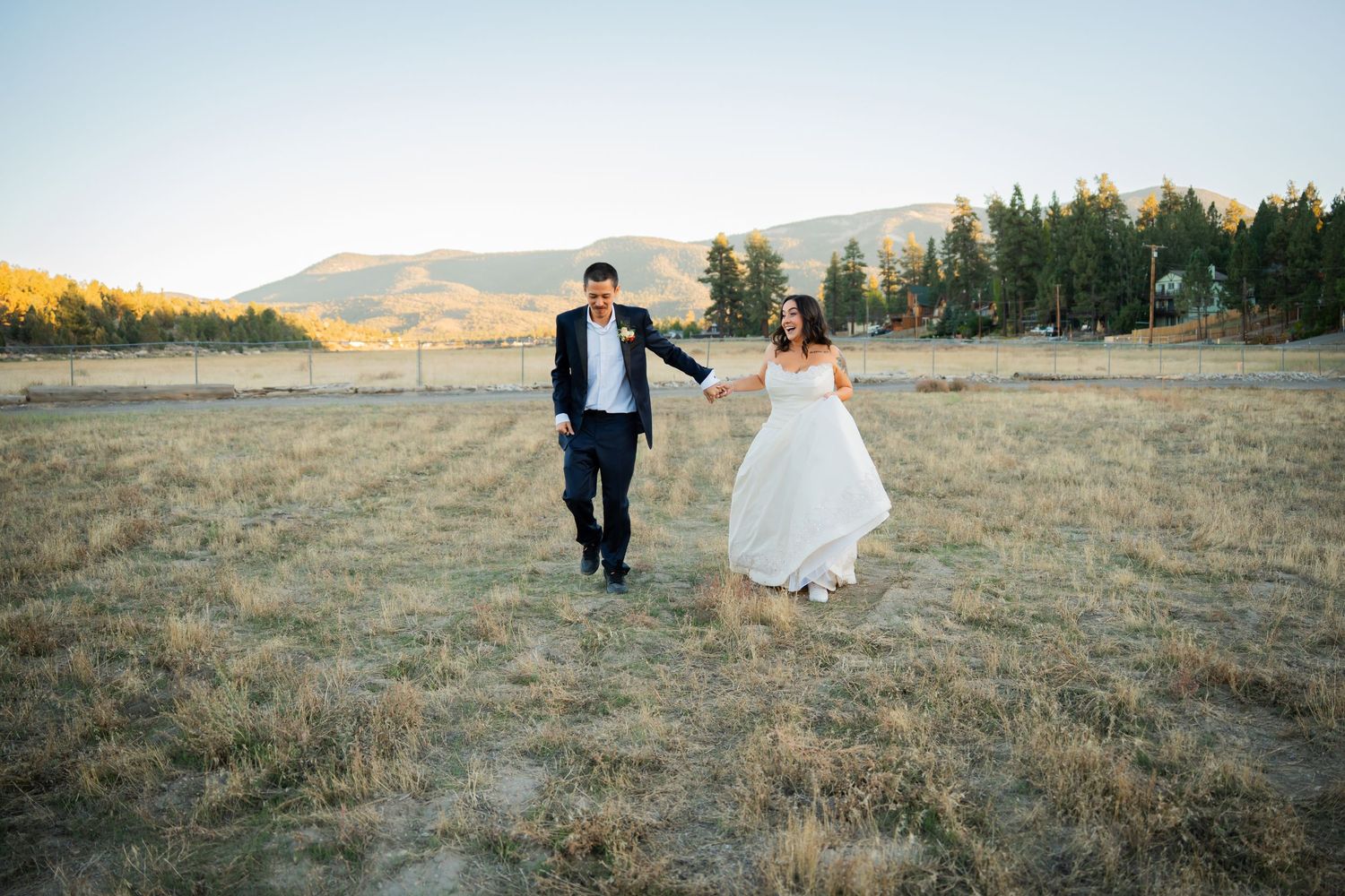 A couple walks together across a golden field at sunset with mountains in the distance.