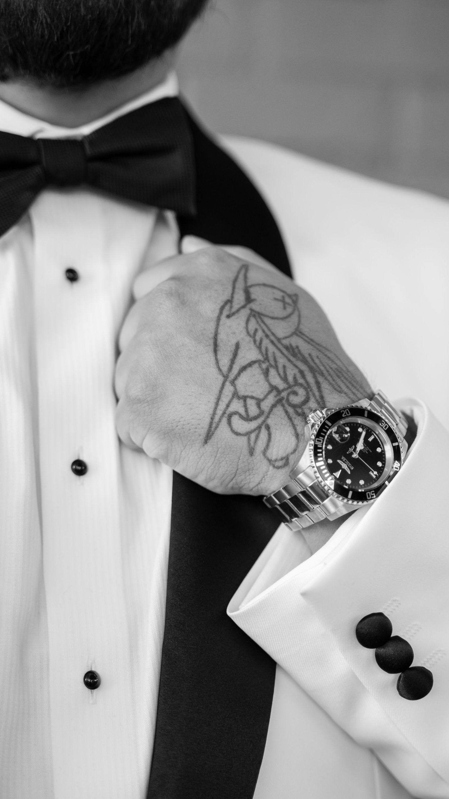 Close-up of groom in tuxedo with black bow tie showing luxury watch and tattoo on hand.