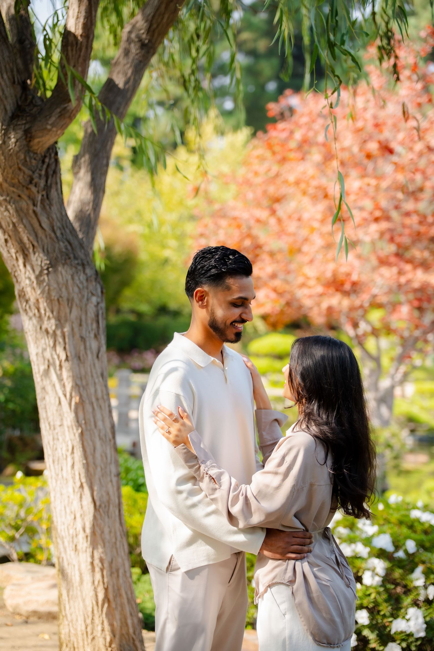Romantic couple in white outfits embracing under autumn trees in a garden setting.