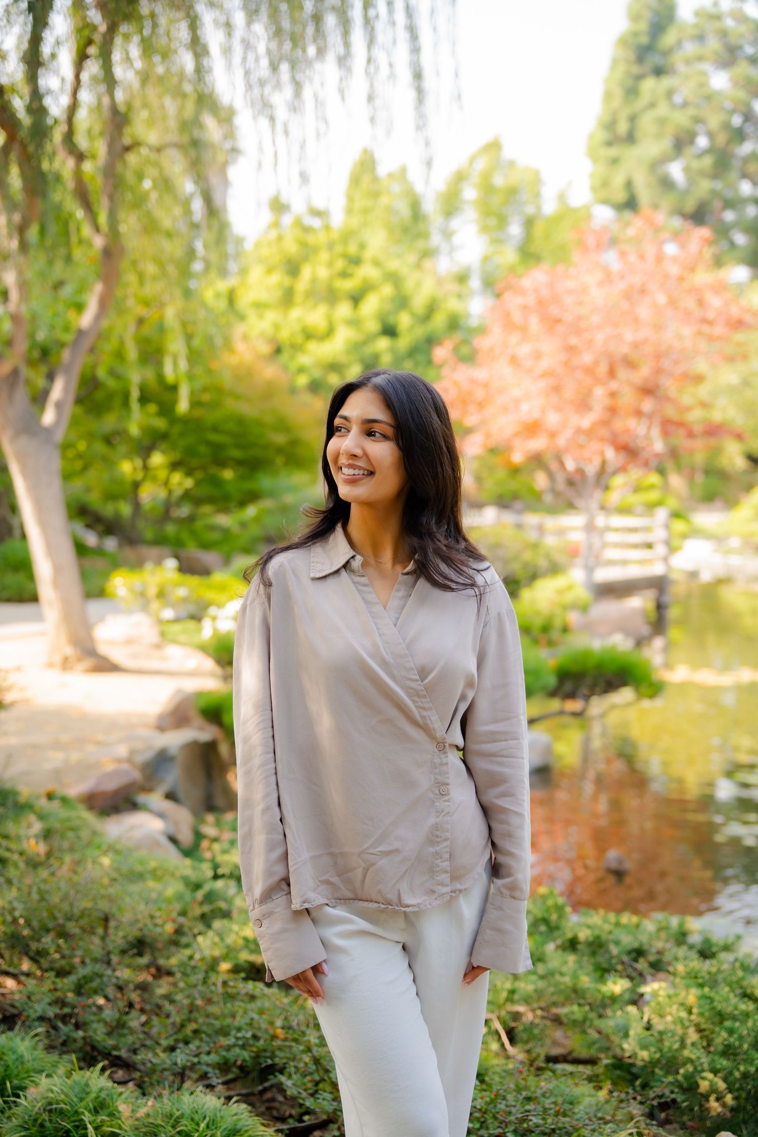 A person in a light gray blazer and white pants poses in a scenic garden setting with willow trees and autumn foliage.