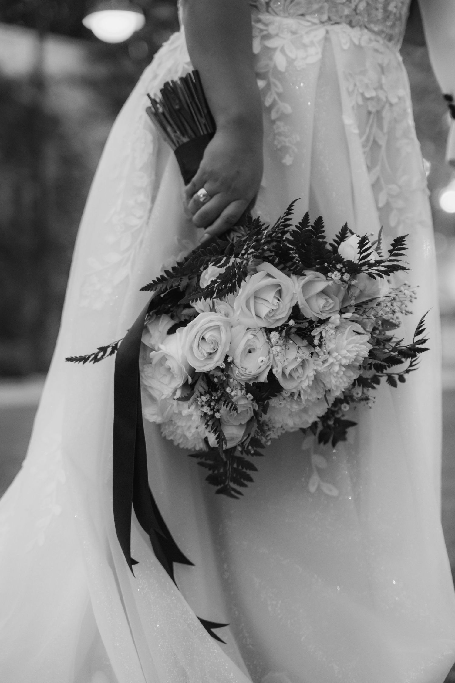 A black and white wedding photo showing a lace dress and elegant bouquet with roses and ferns tied with black ribbon.