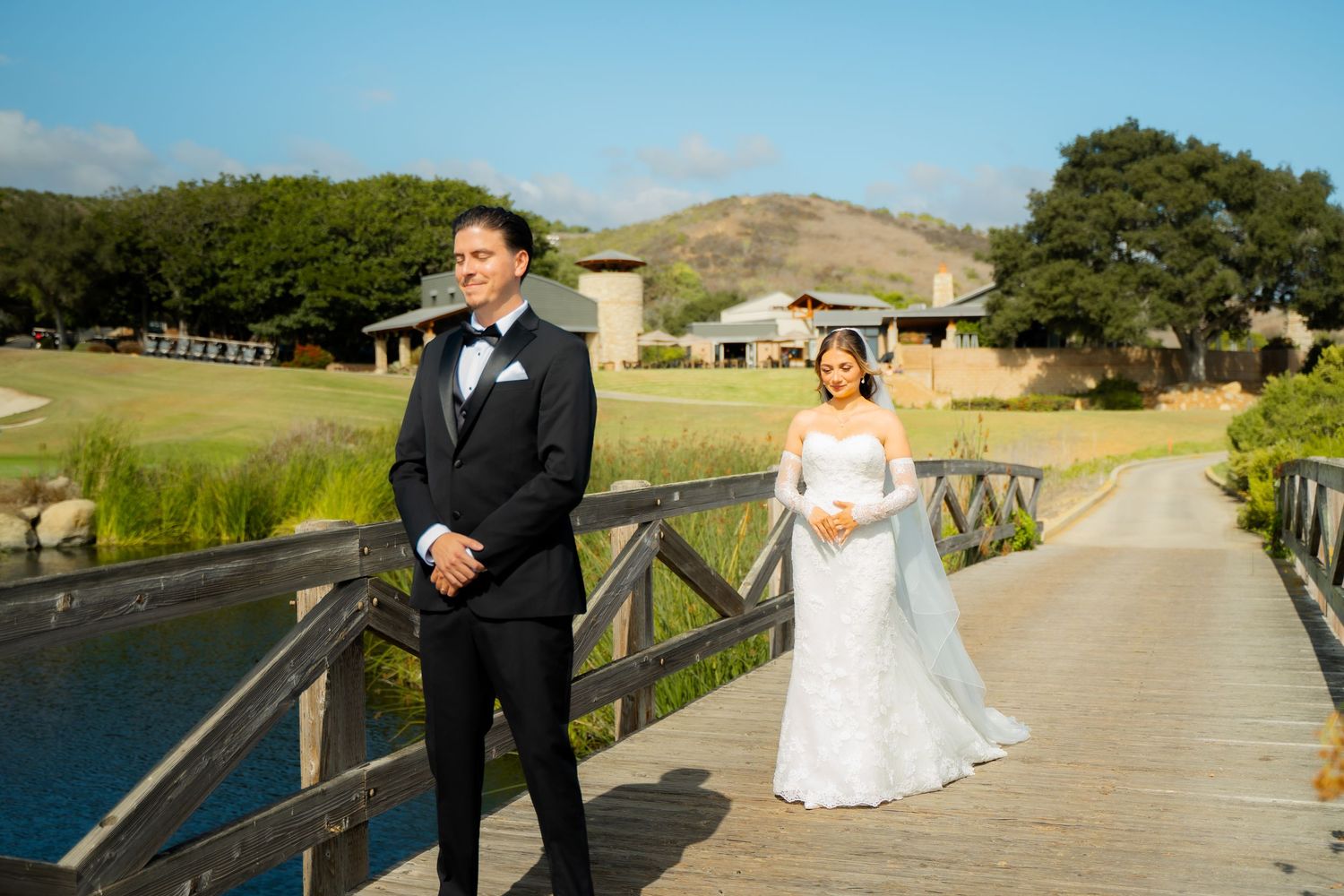 Wedding couple stands on wooden bridge overlooking scenic lake and rolling hills at outdoor venue.
