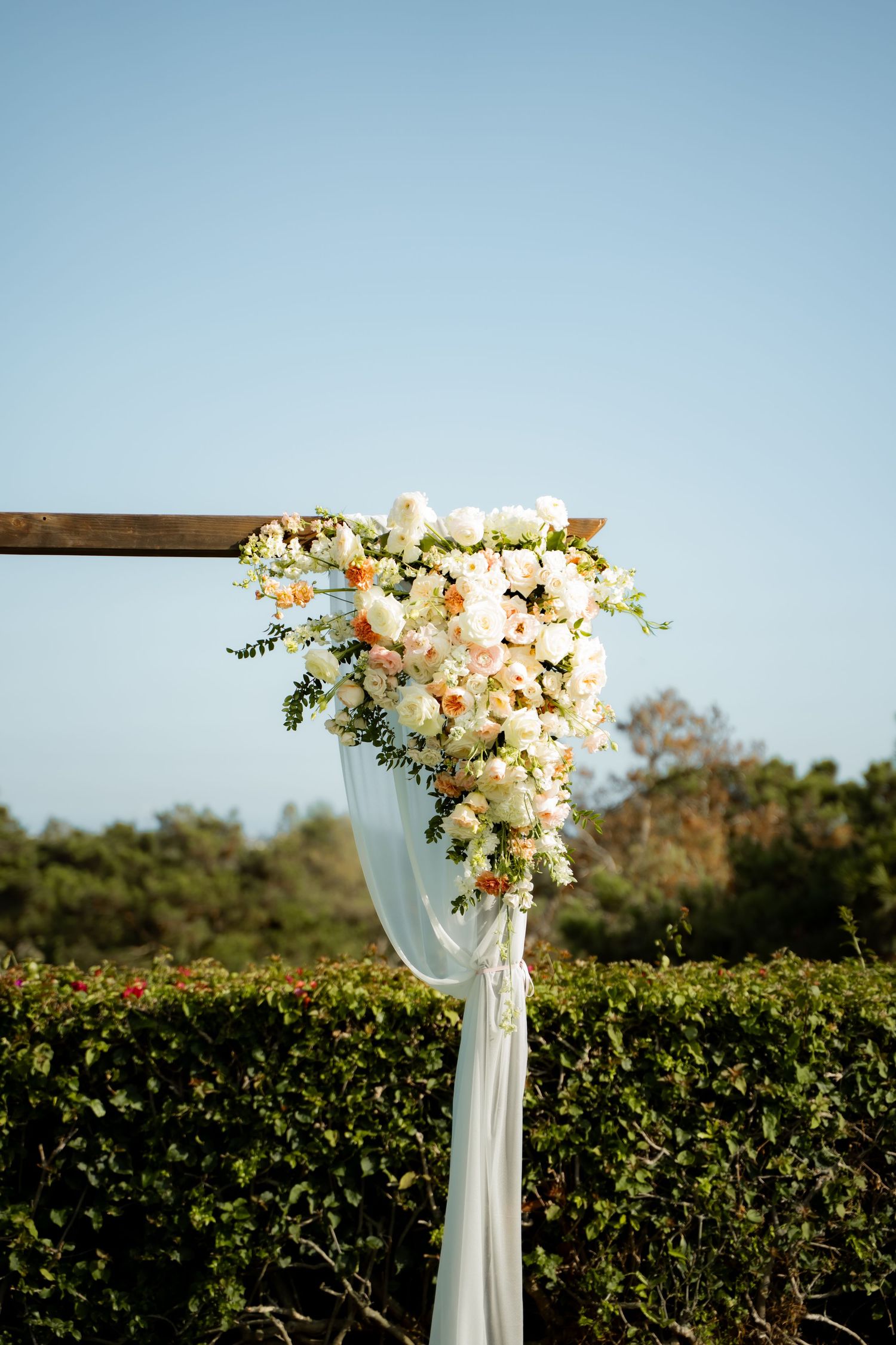 White floral arrangement with draped fabric decorates a wooden beam against a blue sky and hedge backdrop at an outdoor venue.